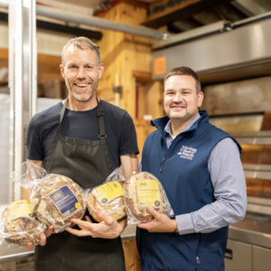 Two men inside of a kitchen holding bread loaves